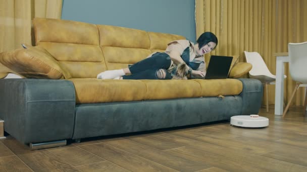 Uma mulher deitada em um sofá e trabalhando em um laptop, e um aspirador de pó robô aspira automaticamente o quarto. Limpeza automatizada de quartos. Tempo livre para trabalhar com seu animal de estimação. - Filmagem, Vídeo