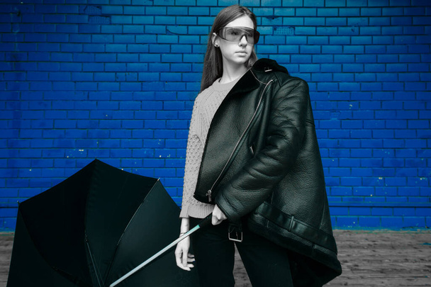 Fashion portrait of artistic woman with umbrella in her hands wearing trendy and popular sheepskin coat and futuristic sunglasses and posing over blu brick wall - 写真・画像