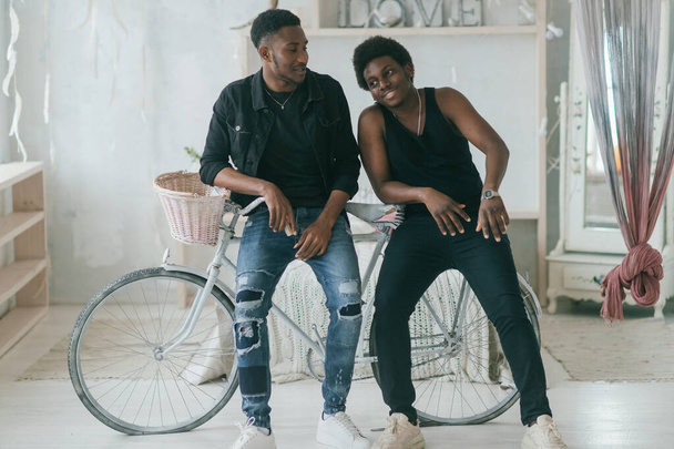 Two happy adult male african frineds standing near bicycle in decorative studio and posing for camera. Two african american men indoor lifestyle vintage style toned portrait - Φωτογραφία, εικόνα