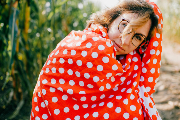 Cheerful young caucasian female with short dark wavy hair in glasses wrapped in red and white coverlet keeps calm in the cornfield in the weekends. - 写真・画像