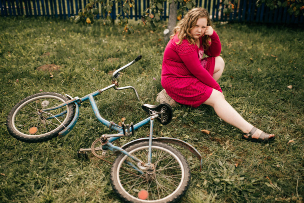 赤いドレスの中髪のハンサムな若い白人の女の子は彼女の古い自転車で庭の草の上に座っており、夏に何かについての夢 - 写真・画像
