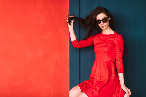 Beautiful fashion girl with long hair, spanish appearance in sunglasses and red elegant dress posing on blue red wall in studio. Amazing babe in suglasses pulling her black hair. Hairstyle advertsing - Photo, image