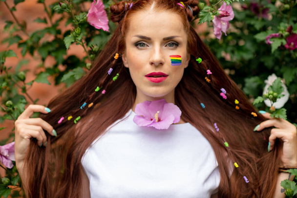 Cheerful lesbian with lgbt rainbow on her face posing in blooming flowers - Valokuva, kuva