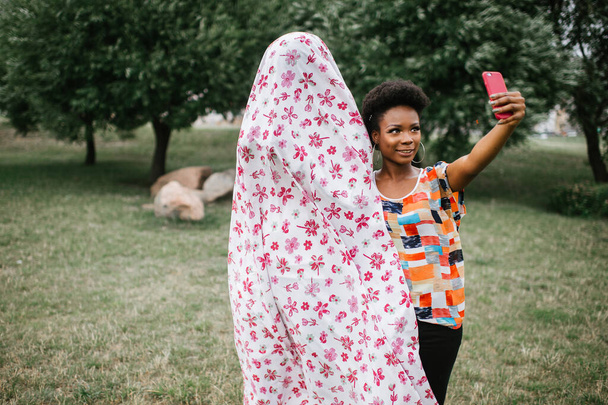 Cheerful african american girl standing with a person in ghost costume outdoor and taking selfie. Halloween party concept. - Photo, image