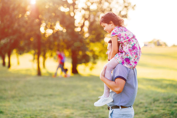 Dad carrying his daughter on shoulders in summer park at sunset. Father have fun with his little child. Kid siiting at parent neck. Happy family playing and making  crazy faces.  Lifestely portrait. - Fotoğraf, Görsel