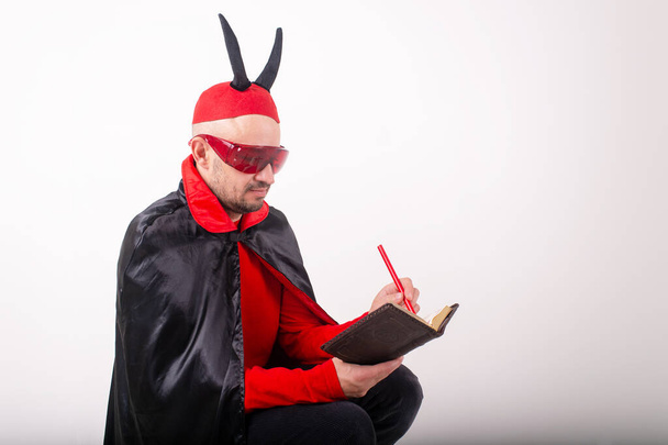Caucasian man in red sunglasses and halloween costume with pen and notebook over white studio background. - Zdjęcie, obraz