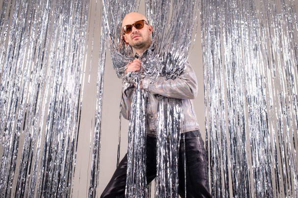 Strange caucasian man in a glittering white shirt, black leather pants, grey shoes and black sunglasses fooling around shiny silver tinsel. - Photo, Image