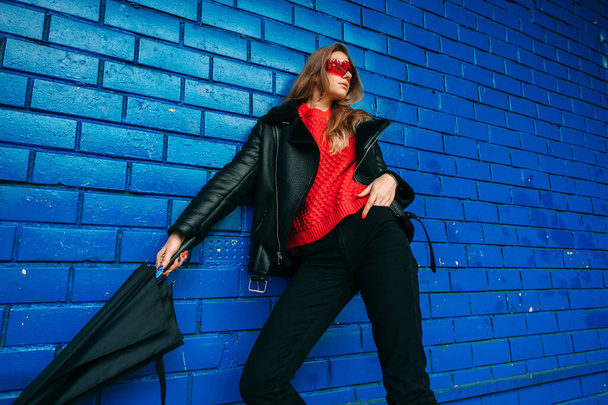 Fashion portrait of young brunette girl in trendy sheepskin caost and red sunglasses posing with umbrella in her hand against blue brick wall outdoors. - Fotoğraf, Görsel