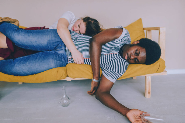 Funny blurred soft focus portrait of odd interracial drunk couple sleeping on bed at home after party. Dark skinned nigerian man lying on bed with caucasian girlfriend and one hand hanging down. - Zdjęcie, obraz