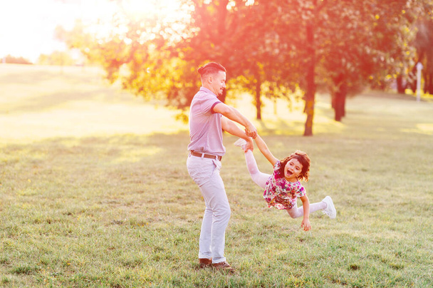 Father holding his daaughter hand with leg and whirling around. Excited family have fun in summer park. Dad playing with little female child on green meadow. Carefree happy childhood. Fatherhood  - Foto, Bild
