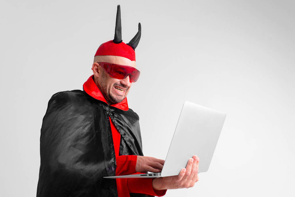 Stylish caucasian man in devil hat with horns and vampire cape with laptop isolated on white backhround. - Foto, afbeelding