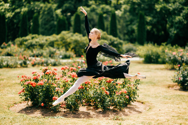 Young ballerina in black dress jumping among rose bushes in park - Фото, зображення
