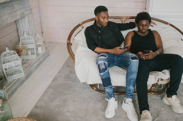 Two happy adult male african frineds sitting  and relaxing together in rattan chair in decorative studio zone. Two african american men indoor lifestyle vintage style toned portrait - Φωτογραφία, εικόνα