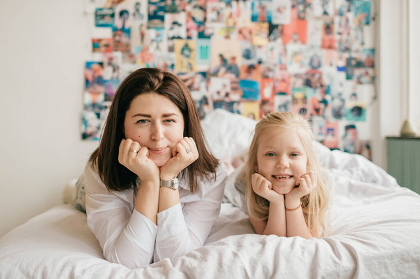 Lifestyle soft focus portrait of happy mom hugs her adorable daughter on white bed. Indoor happy family portrait of smiling mother and her daughter lying together on bed with decorative wall behind - Photo, image