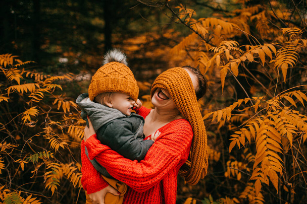Handsome caucasian mother in warm grey jacket, black trousers and grey trainers hugs and rejoices with her little son in warm bright clothes. - Fotografie, Obrázek