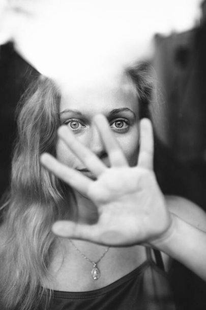 Black-and-white portrait of lovely caucasian young girl with blue eyes, fair hair, pretty face behind glass and covers face with her hand and say to stop - Fotó, kép