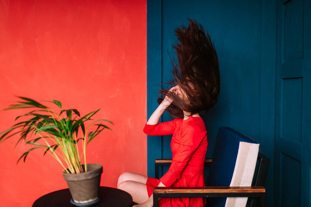 Beautiful fashion girl with black long hair, in sunglasses, red elegant dress sitting in chair on blue red wall in studio. Stylish brunnetre with scattered flying hair. Hairdresser services concept - Foto, Imagem