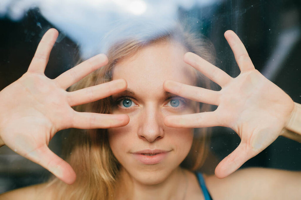 Портрет милой белой молодой девушки с голубыми глазами, светлыми волосами, красивое лицо за стеклом и закрывает лицо руками - Фото, изображение