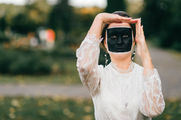 Strange woman in wedding dress with creative face art (black square)showing gestures by her hands in summer park. - Foto, afbeelding