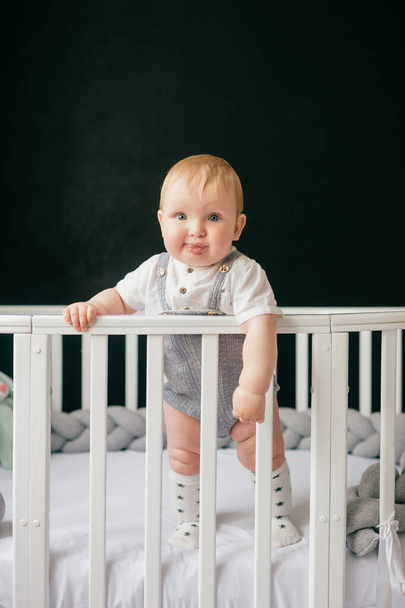 Portrait of funny baby standing in crib. Stylish baby boy standing alone in cot over black background. - Fotografie, Obrázek