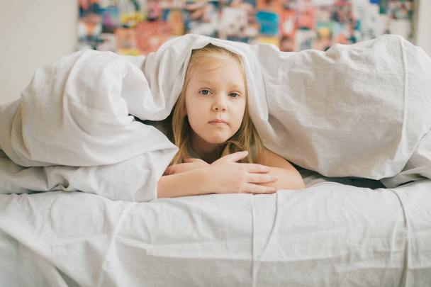 Young adorable blonde girl with funny face lying on bed under white blanket and look at camera with sad eyes. Little female child with pensive face indoor lifestyle portrait. Little  girl portrait - 写真・画像