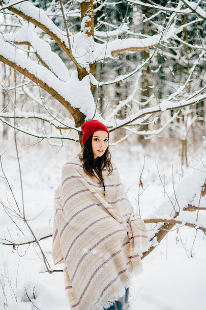 Young attractive girl covering with a warm cape posing among snow branches in the forest - Foto, Imagem