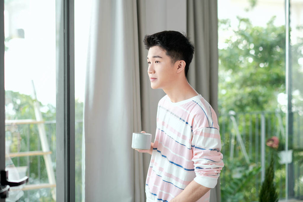 Fiatal jóképű gondtalan férfi közel modern teljes hosszúságú ablak élvezi egy csésze kávét, miközben néz ki - Fotó, kép
