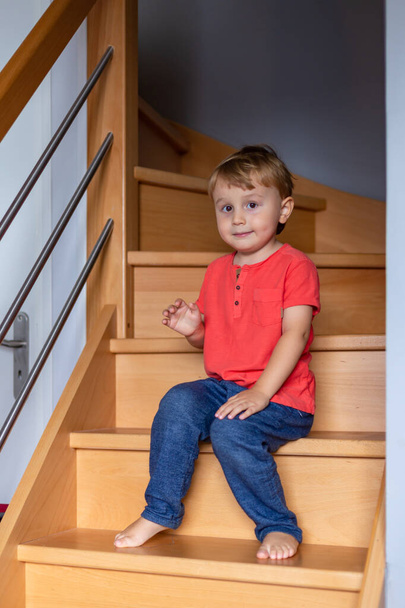 Kleine jongen op blote voeten draagt t-shirt zit thuis op de trap.  - Foto, afbeelding