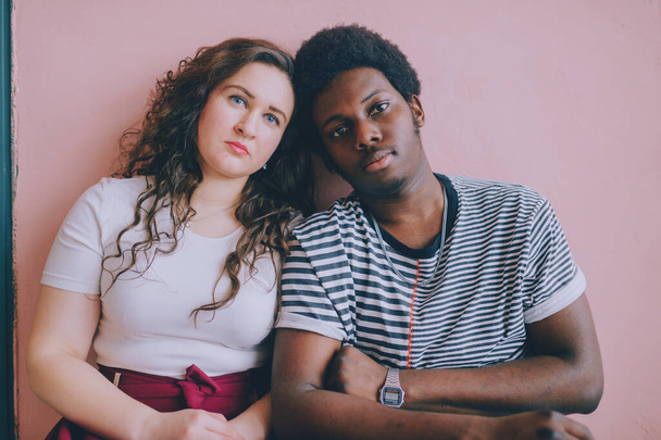 Hugging interracial couple lifestyle indoor portrait on pink wall background. Dark skinned nigerian man in striped shirt embracing caucasian girlfriend in red dress at home. International friendship - Фото, изображение