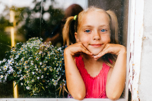 Μικρό χαριτωμένο καυκάσιο αυτο-απομονωμένο κορίτσι με μακριά ξανθά μαλλιά και με ροζ φόρεμα κρατά λουλούδια και χαμόγελα πίσω από το παράθυρο κατά τη διάρκεια πανδημίας του coronavirus covid 19. Μείνε σπίτι. Κοινωνική υστέρηση. - Φωτογραφία, εικόνα