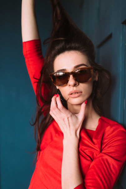 Beautiful fashion girl with dark long hair, spanish appearance in sunglasses and red elegant dress posing on blue wall in studio. Indoor lifestyle portrait of stylish woman. Aamzing brunette model - Zdjęcie, obraz