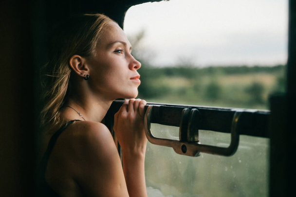 Mood atmospheric lifestyle portrait of young beautiful blonde hair girl looking out of window from riding train. Pretty teen enjoying beauty of nature from moving train car in summer. Travel concept. - Foto, imagen