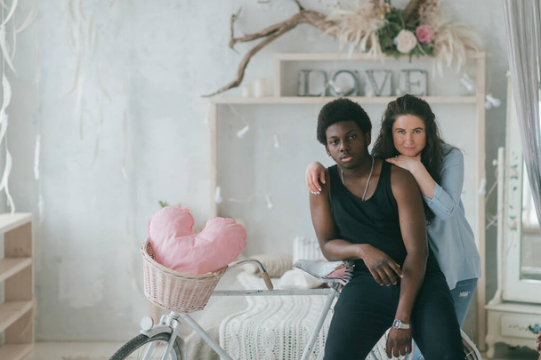 Multicultural love and relationships concept. Loving and embracing interracial couple indoor vintage style portrait. Dark skinned man standing near bicycle with his cheerful happy white girlfriend - Photo, image