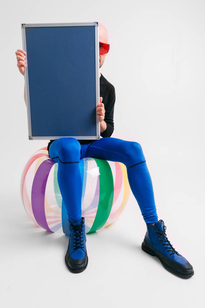 Stylish coach dressed in blue tights sitting on beach ball and holding nameplate in his hands over white background - Foto, imagen