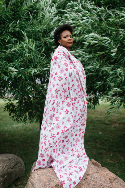 Beautiful dark skinned girl wrapped in sheet with fashion print posing at park with trees on background. - Zdjęcie, obraz