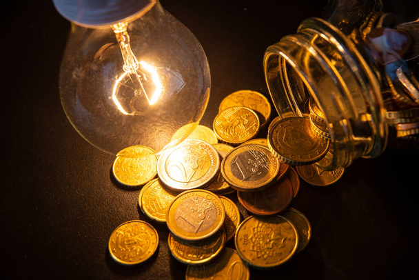 Γυάλινο βάζο με κέρματα και λάμπα δίπλα του. Εξοικονόμηση λογαριασμών κοινής ωφέλειας. Κόστος ηλεκτρικής ενέργειας - Φωτογραφία, εικόνα