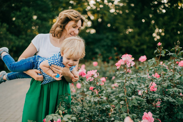 Beautiful caucasian mum with dark hair in white t-shirt and long green skirt plays in the park with her lovely son with short fair hair in white and blue shirt and blue jeans. - Fotoğraf, Görsel