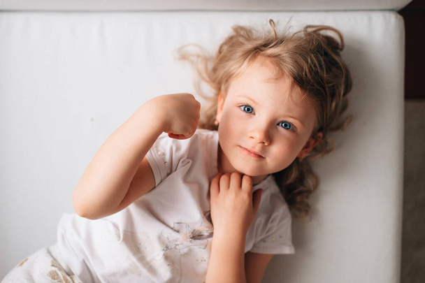 Portrait of cute little caucasian girl in white sleepwear lies on her white bed and rejoices - Foto, Imagem