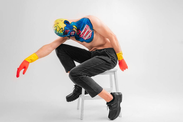 Portrait of a strange man with makeup on his head and shoulder in red-yellow gloves, black pants and boots, no T-shirt sits on a white chair - Фото, зображення
