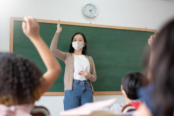 Aasian opettaja yllään suojaava kasvonaamio ja Lapset tai Schoolkids nostaa kätensä luokkahuoneessa, kun taas covid-19 pandemia. Uusi normaalin elämän käsite. - Valokuva, kuva