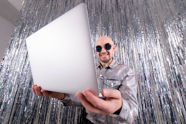 Busy caucasian businessman without hair in silver shirt, black leather trousers and grey trainers with black sunglasses do freelance with his laptop on silver tinsel background. - Fotoğraf, Görsel