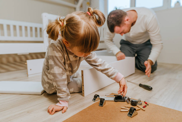 Pequeña niña de 4 años ayuda a su padre a montar o arreglar el cajón de la cama en el dormitorio de los niños. - Foto, Imagen