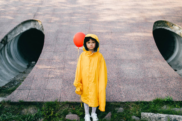 Portrait of pretty litle girl in big adult oversized yellow raincoat with red balloon in hand standing and sitting against sewage tunnel pipe. Horror movie concept. Funny child posing in summer day - Fotoğraf, Görsel