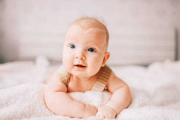 Adorable lovely newborn male baby boy with smiling emotional  happy face lifestyle indoor portrait. Funny infant child lying on stomach on bed with white wall on background.  Carefree childhood - Fotó, kép