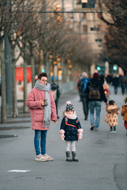 Picture of young beautiful caucasian mum with her cheerful daughter go for a walk on the promenade in autumn or spring - Photo, Image