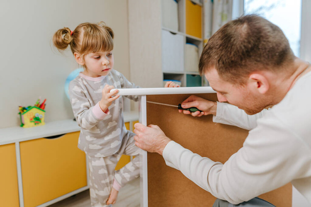 Little 4-years girl helps her father assemble or fixing the drawer of bed in the kids bedroom. - Zdjęcie, obraz