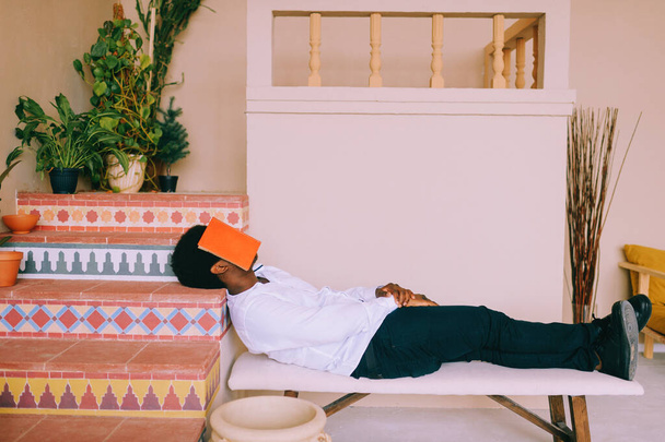 Extraño retrato de lyfestyle de un joven africano de piel oscura acostado en el banco y durmiendo con un libro en la cara. Cansado después de leer extraño negro nigeriano niño descansando en casa con libro rojo cubrir su cara - Foto, Imagen