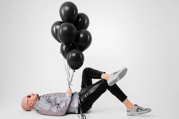 Beautiful caucasian man without hair in grey shirt, black leather trousers and grey trainers with black sunglasses lies on the floor and holds black baloons isolated on white background. - Foto, immagini