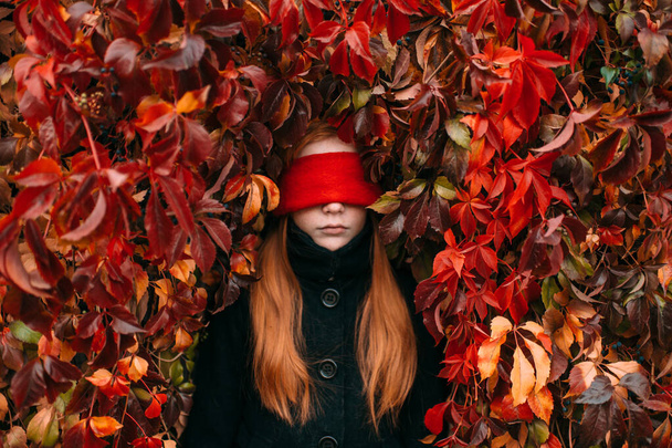 Portrait of longhaired redhead girl with red blindfold in her eyes standing against ivy wall in autumn park. - Фото, изображение