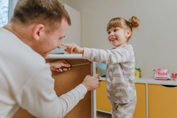 Маленька 4-річна дівчинка грає зі своїм батьком, коли він збирає ящик ліжка в дитячій спальні
. - Фото, зображення